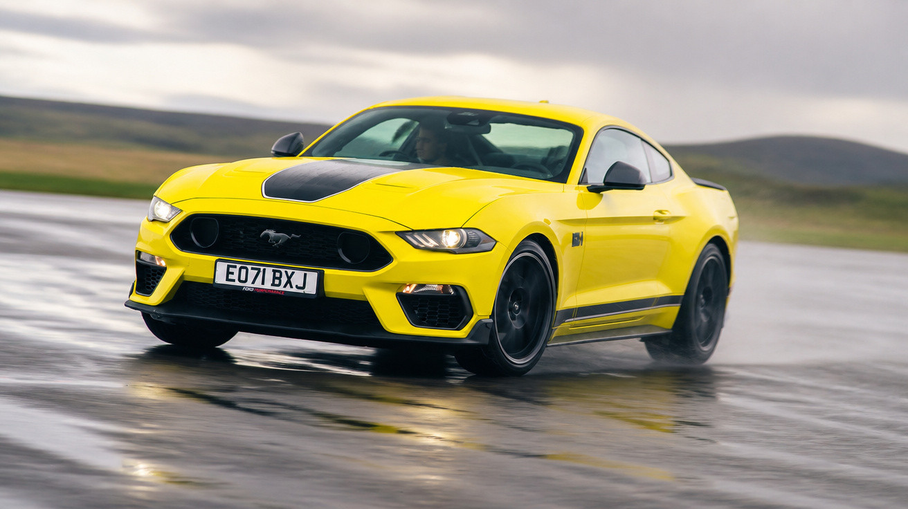 Carro amarelo desportivo Ford Mustang em movimento numa estrada molhada com fundo natural desfocado.