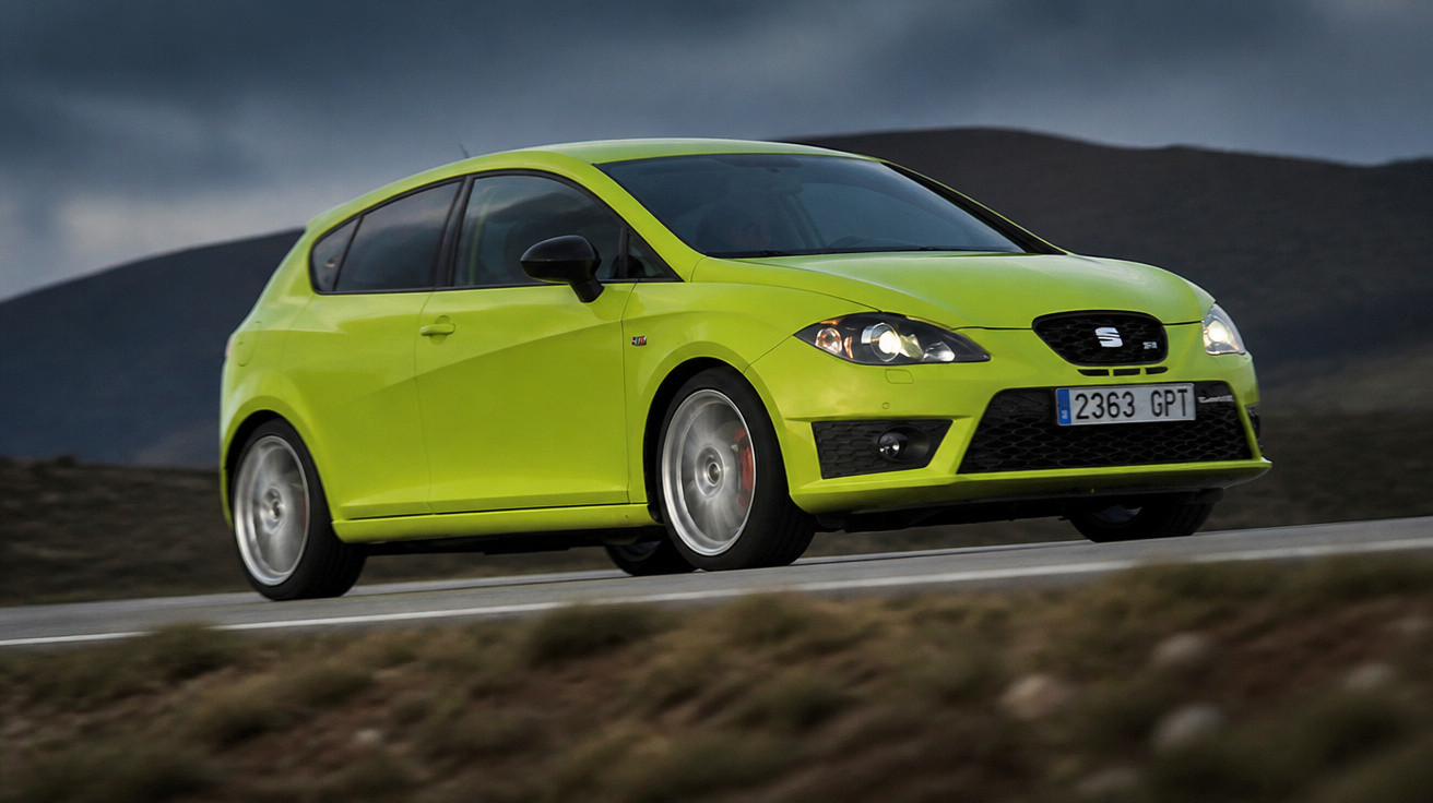 Carro Seat Leon amarelo em estrada com céu nublado e montanhas ao fundo em movimento.