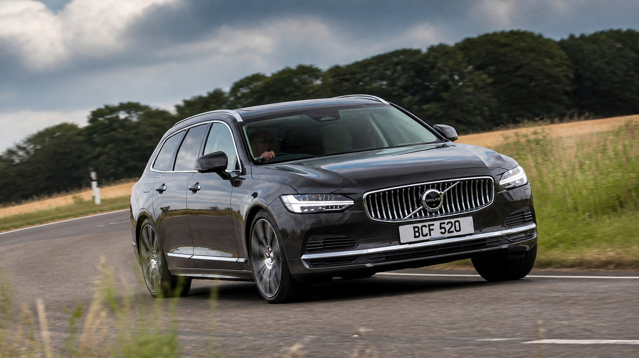 Carro Volvo preto a circular numa estrada curva rodeada de vegetação e céu nublado.