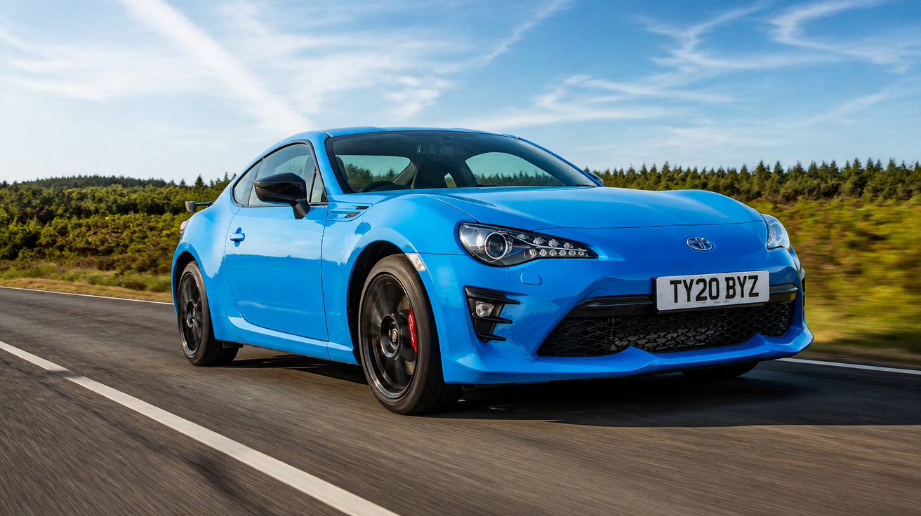 Carro desportivo azul a alta velocidade numa estrada rodeada de vegetação e céu azul com nuvens.