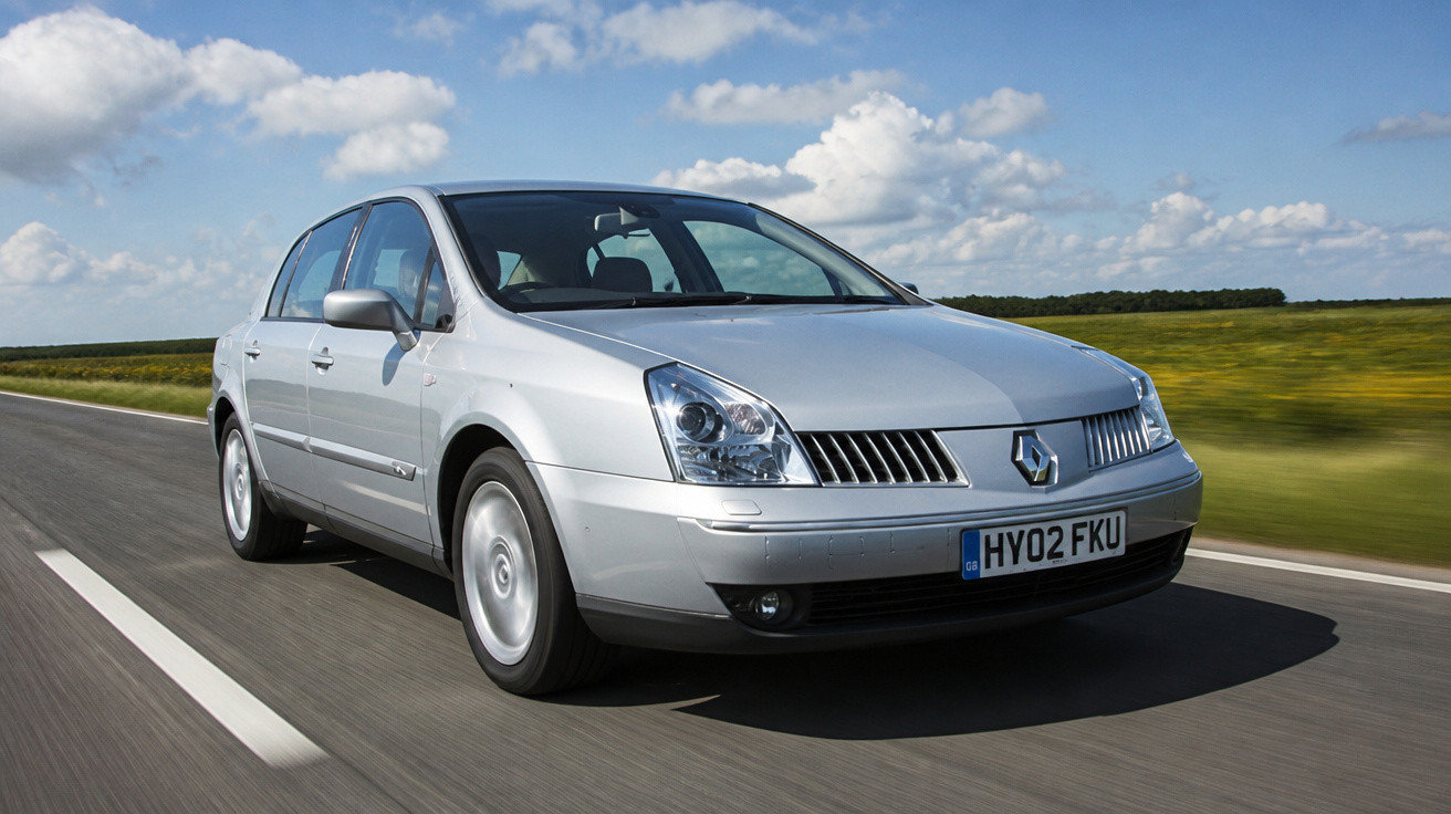 Carro sedan prata a circular numa estrada rodeada de campos verdes e céu azul com nuvens.